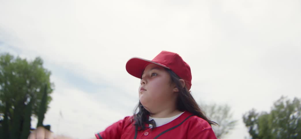 Slow motion, low-angle shot of a determined young girl in a red baseball uniform pitching a ball