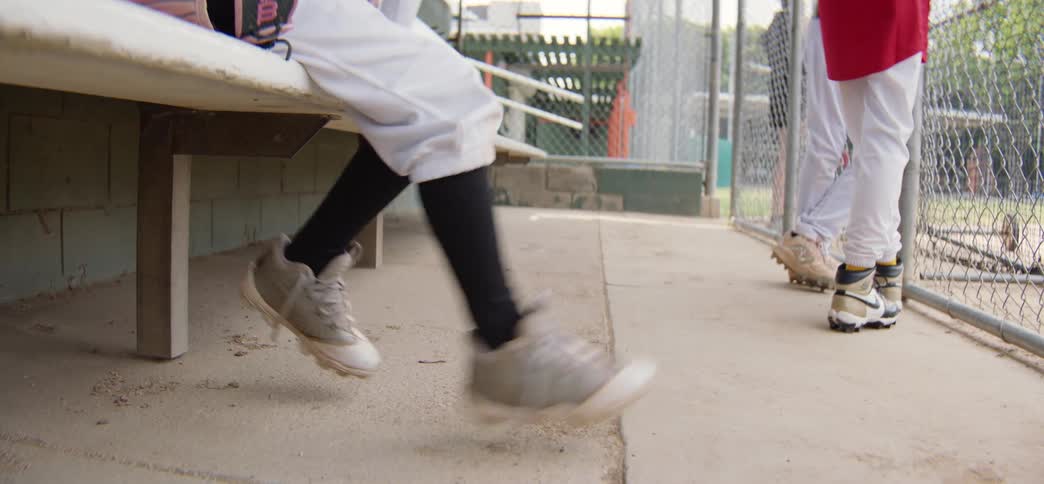 A young baseball player in uniform nervously swings their legs while sitting on the bench in the dugout