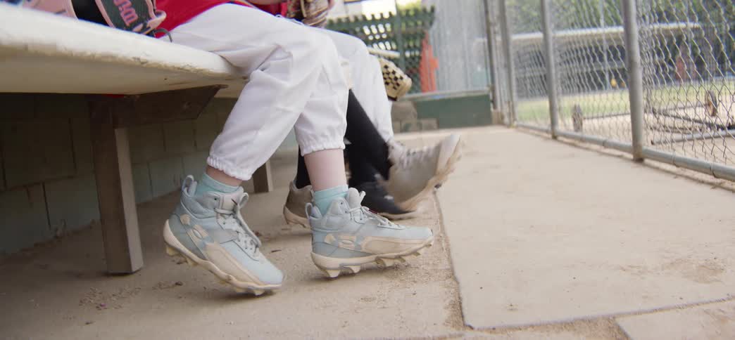 A low angle shot shows the feet of young baseball players swinging their legs while waiting on the bench in the dugout