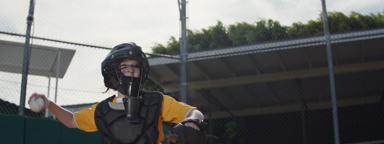 A young baseball catcher in full uniform catches a pitch and throws the ball back in cinematic slow motion