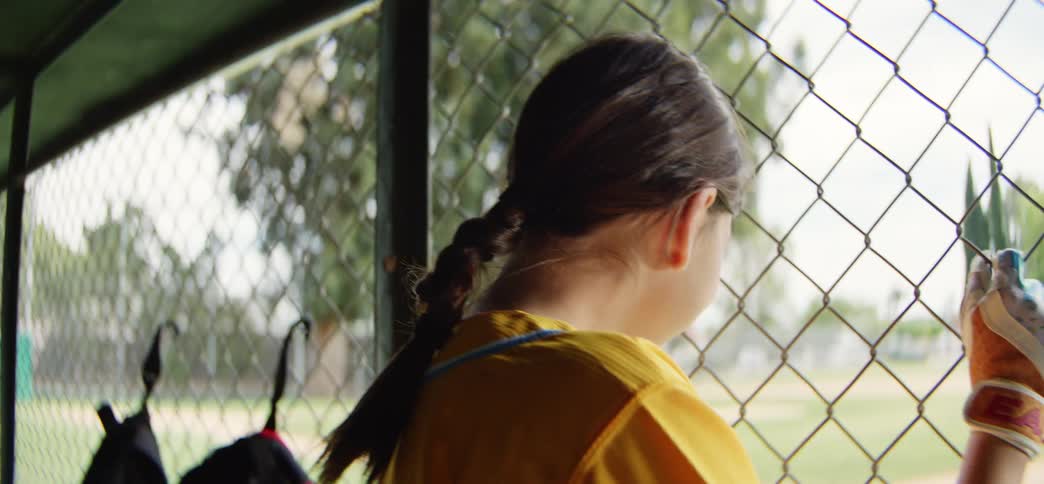 A smiling young girl in a yellow jersey and batting glove watches her softball game from the dugout in slow motion