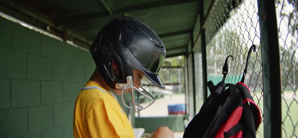 A young baseball player in a yellow uniform puts on his helmet and gloves in the dugout in slow motion