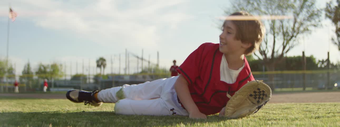 A young boy in a baseball uniform makes a spectacular diving catch across the grass in slow motion on a sunny field