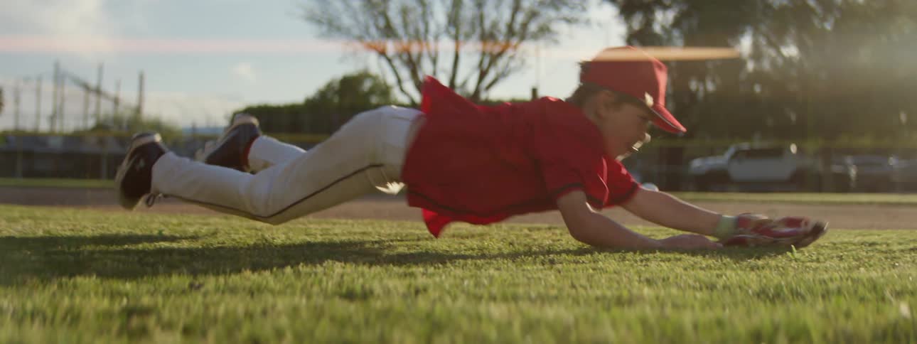 A determined young baseball player dives across the outfield grass to make a spectacular catch in slow motion