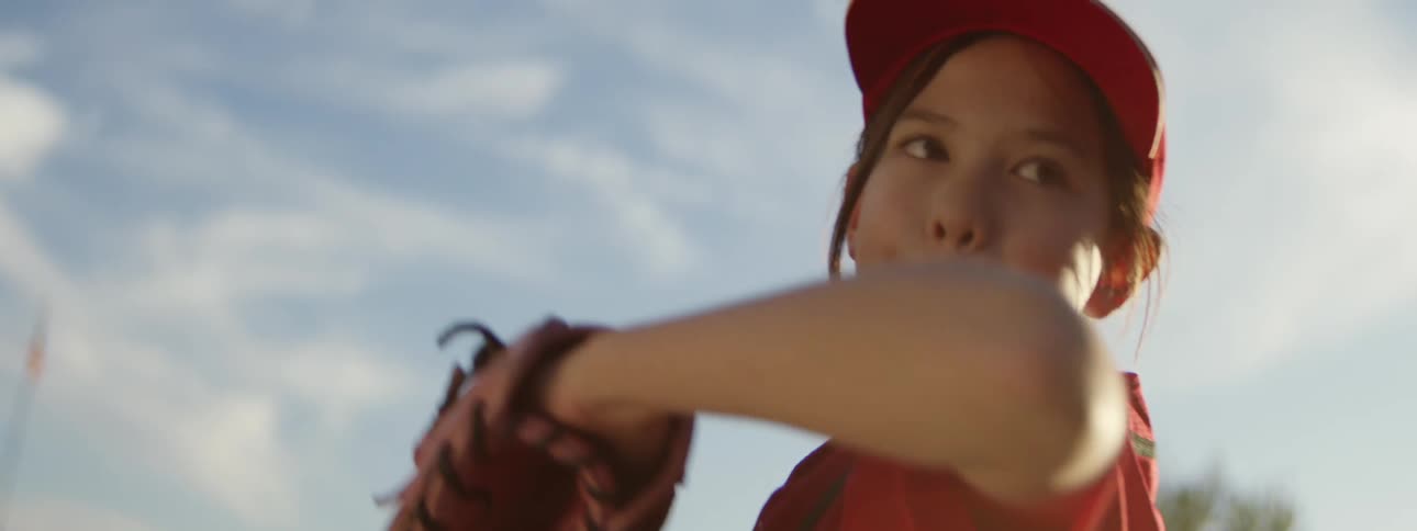 A young girl in a red baseball uniform throws a pitch in slow motion on a sunny day