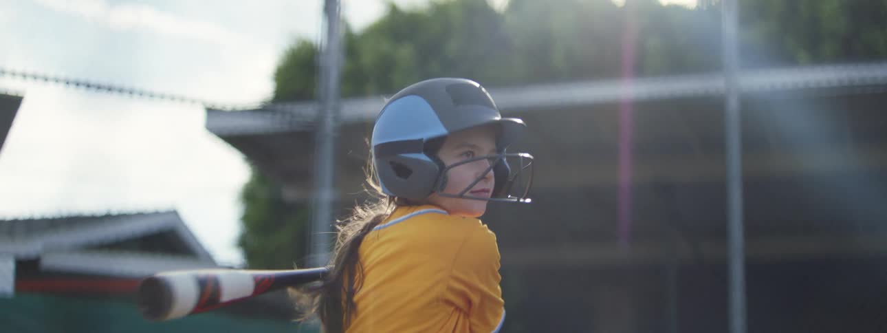 A young girl in a yellow baseball uniform and helmet swings her bat in dramatic slow motion on a sunny day
