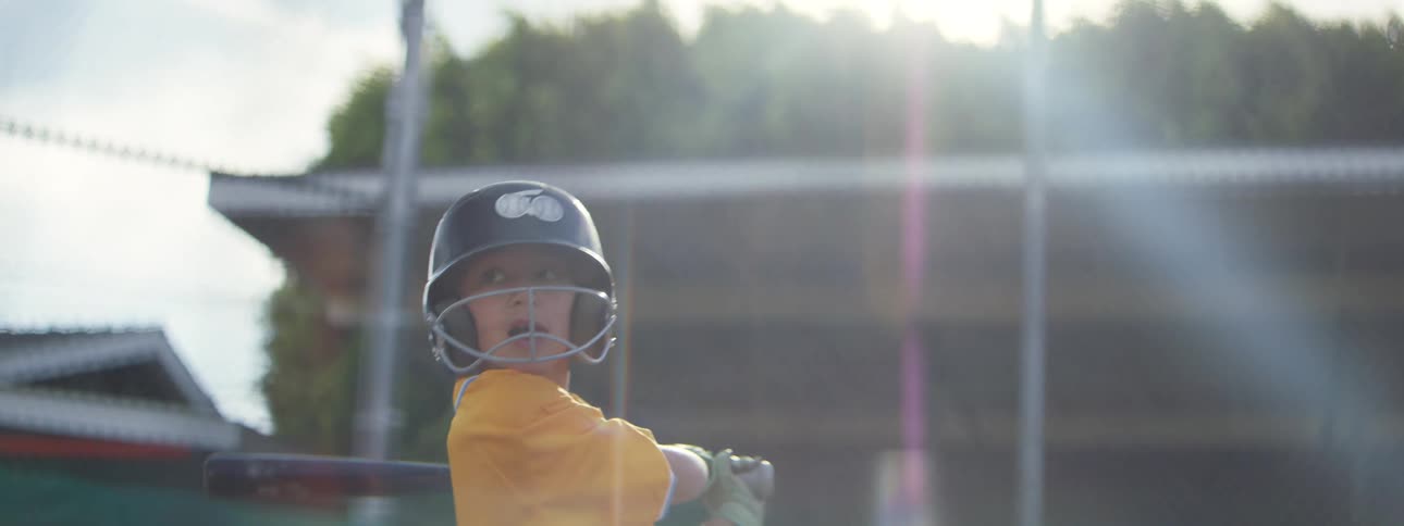 A determined young little league player in a yellow uniform hits a baseball in dramatic slow motion with a sunny lens flare