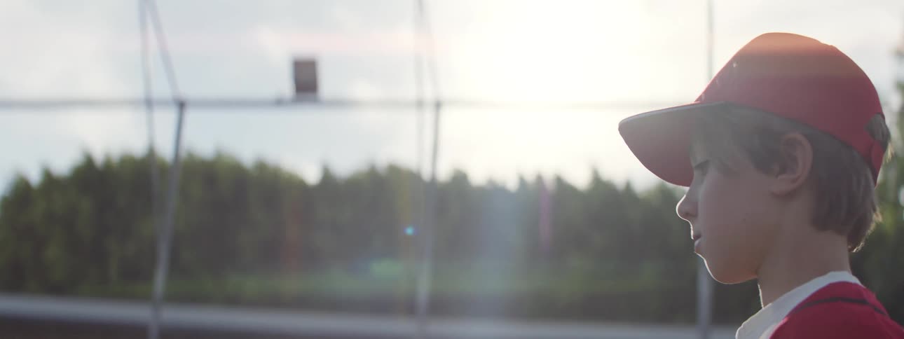 Profile of a determined young baseball player in a red cap tossing a ball into the sunlit sky in slow motion