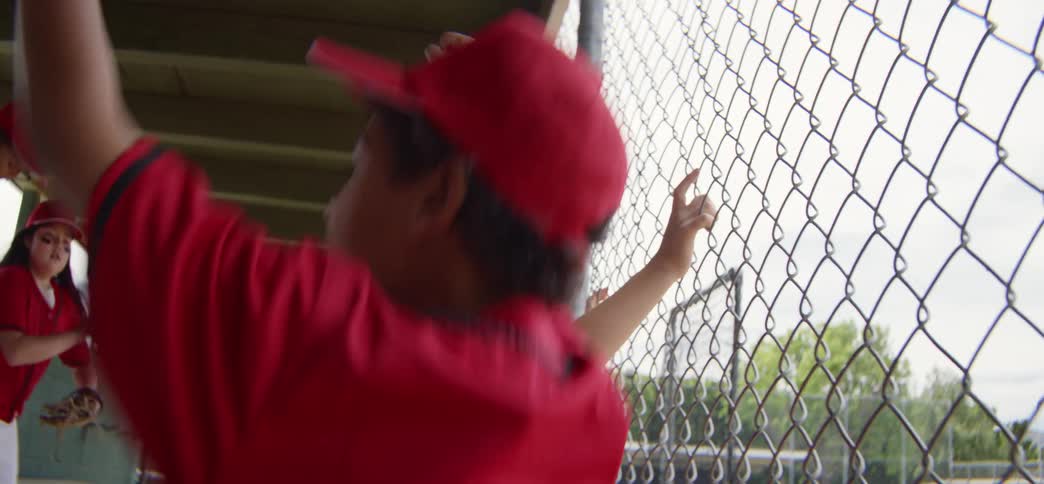 Excited little league players in red uniforms cheer and clap for their teammates from behind the dugout fence
