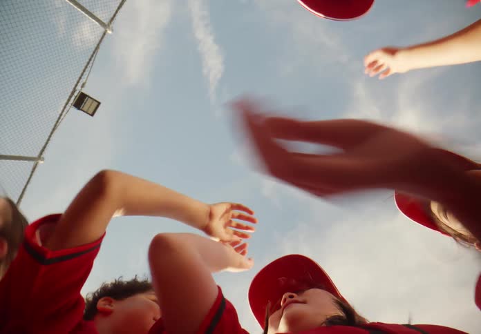 From a low angle, a youth baseball team puts their hands in for a huddle and cheers in celebration against a bright blue sky