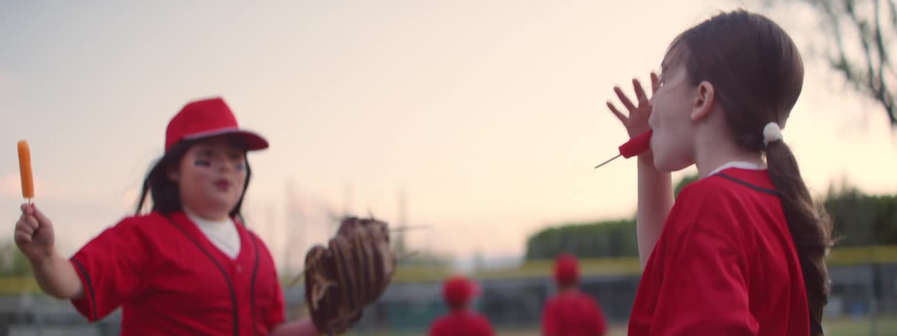 Slow motion side view of two young girls in red baseball uniforms eating popsicles together on a baseball field at sunset