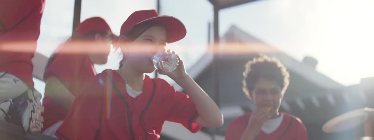 A young girl in a red baseball uniform takes a refreshing water break in the dugout on a sunny day