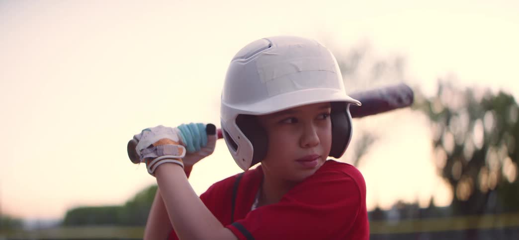 A determined young girl in a batting helmet swings a baseball bat in slow motion at sunset