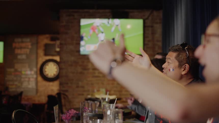 Passionate soccer fans point and cheer in slow motion as they watch their team score on television in a bar