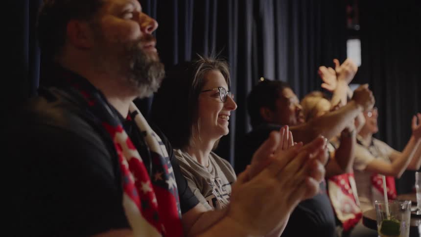A diverse group of excited sports fans cheers and claps in slow motion while watching a game at a bar