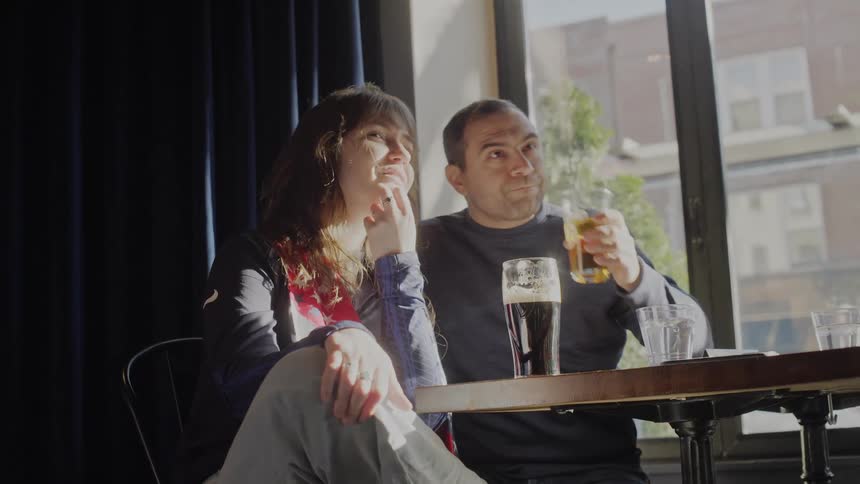 A man and woman enjoy beers and conversation while watching something off-screen at a sunlit table in a pub
