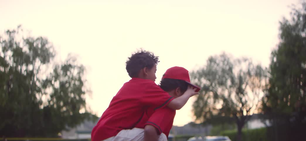 An older brother gives his younger brother a piggyback ride in their baseball uniforms in slow motion