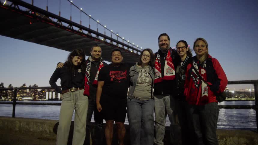 A diverse group of smiling friends wearing team scarves pose for a photo under a large illuminated bridge at twilight