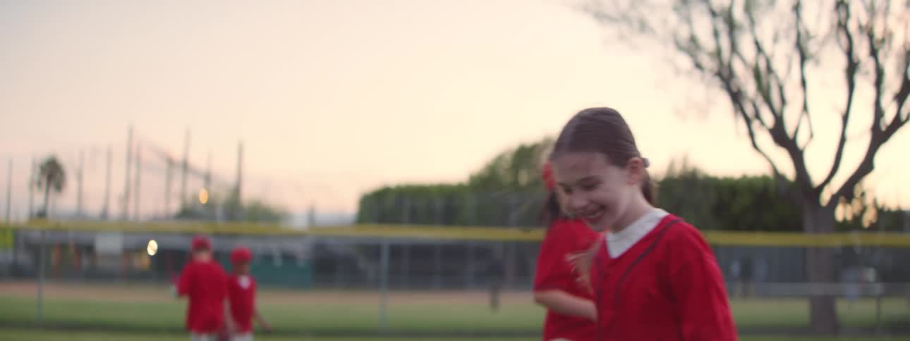 Young baseball players in red uniforms joyfully celebrate with popsicles on the field at sunset in slow motion