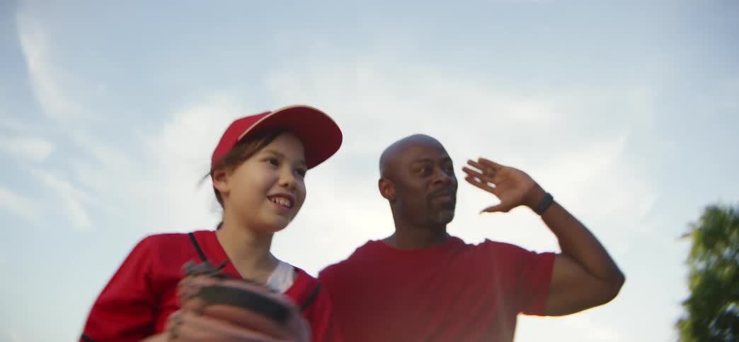 A happy young girl in a red baseball uniform listens as her encouraging coach walks with her onto the field in slow motion