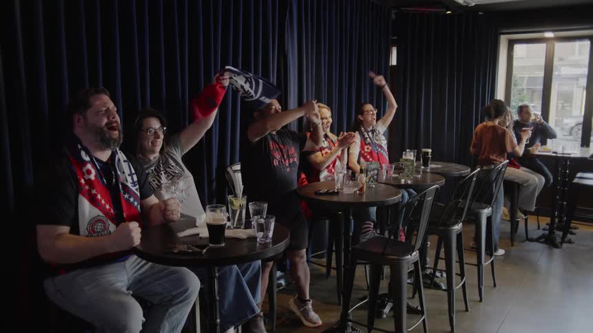 A diverse group of excited sports fans erupt in celebration while watching their team score a goal at a local pub