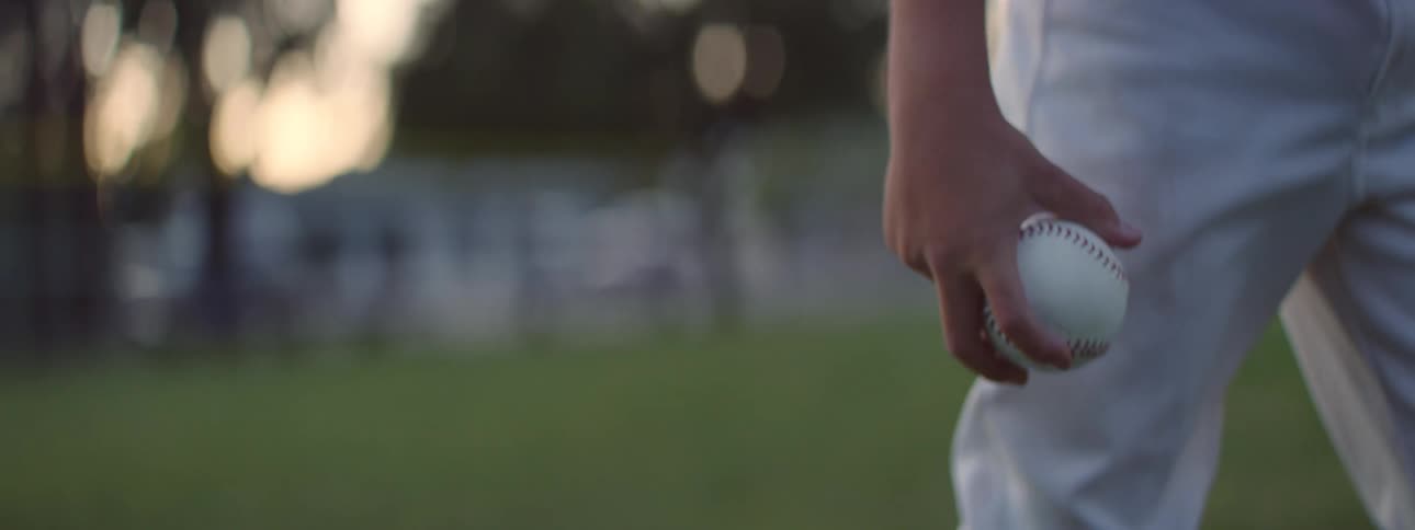 A cinematic slow-motion close-up of a young baseball player's hand holding a baseball on a grassy field at dusk