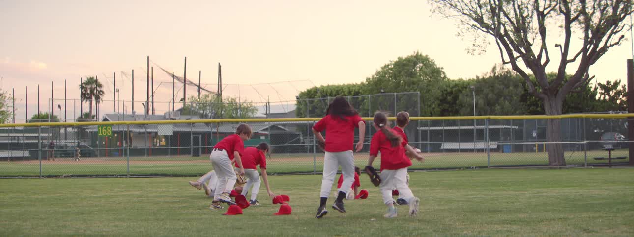 A team of diverse young baseball players in red uniforms celebrate a victory by throwing their hats in the air and tackling each other in slow motion