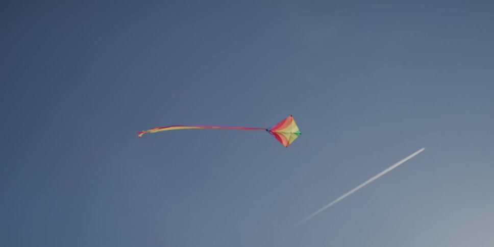 A colorful kite flies serenely in a clear blue sky with a jet contrail visible in the distance