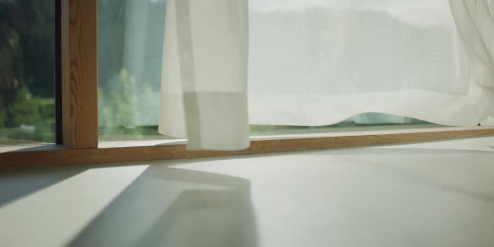 A gentle breeze blows through sheer white curtains in slow motion, with sunlight casting dancing shadows on the floor
