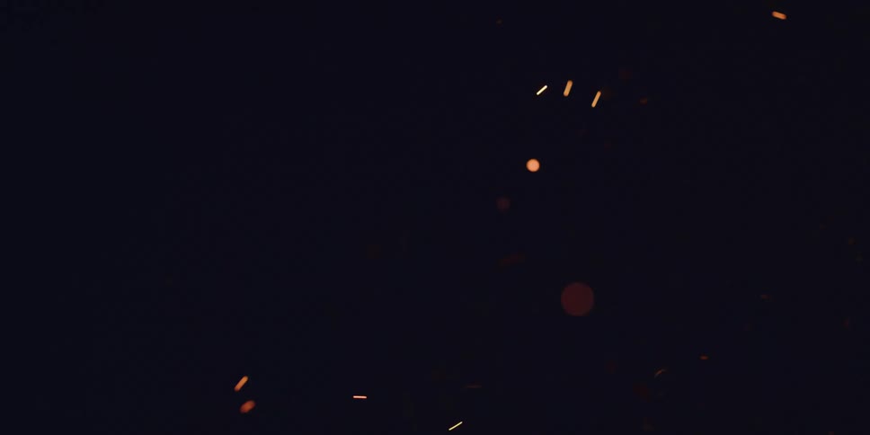 Glowing embers rise from a bonfire in slow motion, creating a mesmerizing bokeh effect against the dark night sky