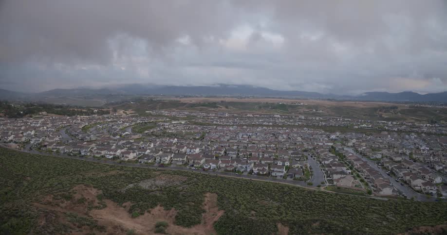 A slow aerial push-in shot reveals a large suburban housing development next to a natural, undeveloped hillside on an overcast day