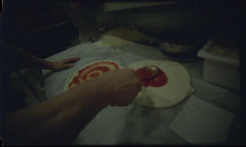 Vintage Super 8 footage captures a chef spreading tomato sauce in a spiral motion on two pizza doughs in a restaurant kitchen