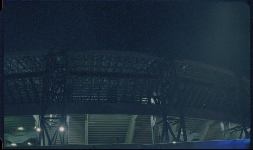 A grainy, vintage film shot of the exterior of a large, empty sports stadium illuminated at night