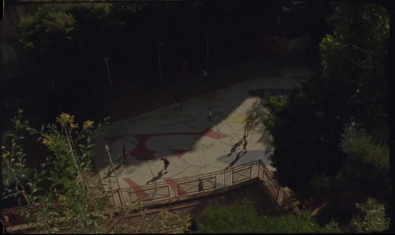 A high-angle vintage film shot captures kids playing basketball on a colorfully painted court in the afternoon sun