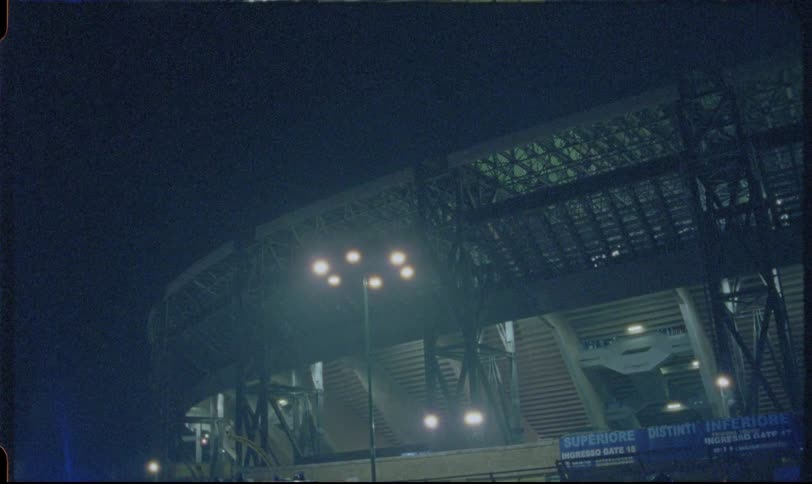 Vintage film-style footage shows the exterior of a massive sports stadium at night with police cars guarding the entrance