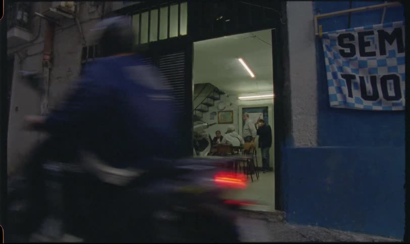 A glimpse into a brightly lit shop from a dark alleyway where men socialize as a scooter drives by