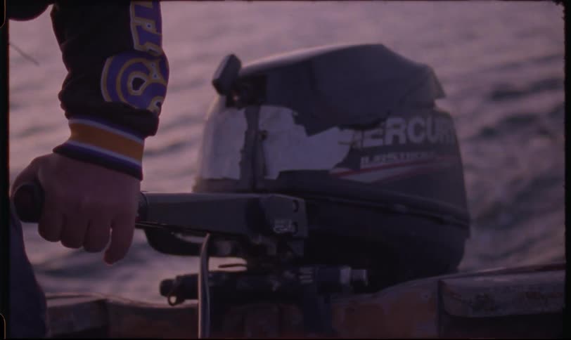A vintage, close-up shot of a hand steering a small boat's outboard motor across the water at dusk