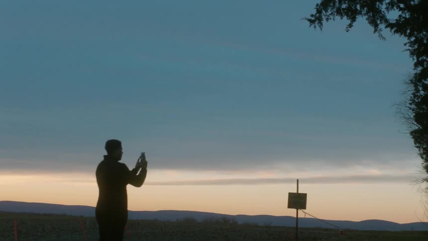A man takes a picture of a sunset in a rural field