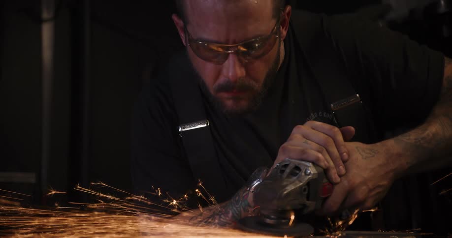A metalworker with safety glasses intently grinds a piece of metal, sending a trail of hot sparks flying