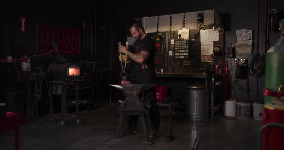 A blacksmith with tattoos works on a piece of hot metal with tongs and a file at his anvil in a dimly lit workshop