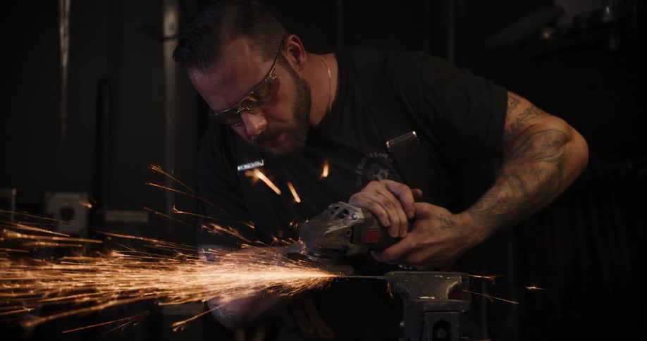 A tattooed craftsman in a dark workshop uses an angle grinder on a piece of metal, creating a dramatic shower of sparks in slow motion