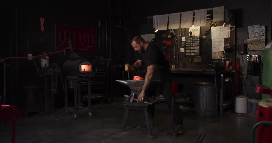 A tattooed blacksmith removes a glowing hot piece of metal from a forge and begins hammering it on an anvil in his dimly lit workshop