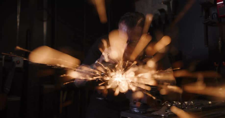 A tattooed craftsman grinds metal in his dark workshop, with a spectacular shower of sparks flying towards the camera