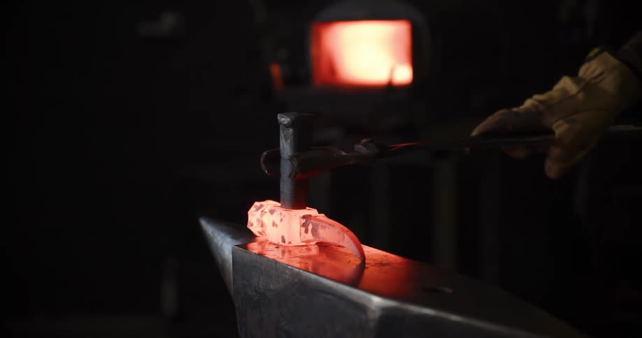 A blacksmith pulls a glowing hot piece of metal from a forge and hammers it into shape on an anvil in a dark workshop