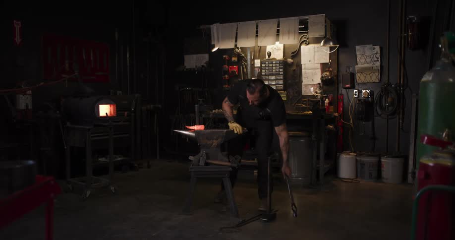 A skilled blacksmith hammers a piece of glowing hot metal on an anvil in his dimly lit workshop