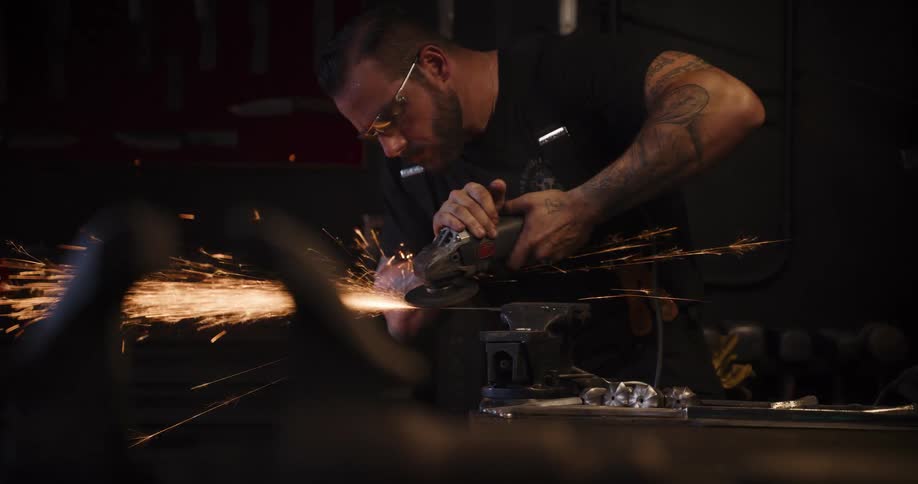 A tattooed craftsman in safety glasses uses an angle grinder in his workshop, creating a shower of sparks in slow motion