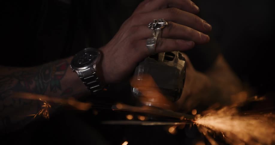 A tattooed craftsman uses an angle grinder to shape a piece of metal, sending a cascade of fiery sparks flying in a dark workshop