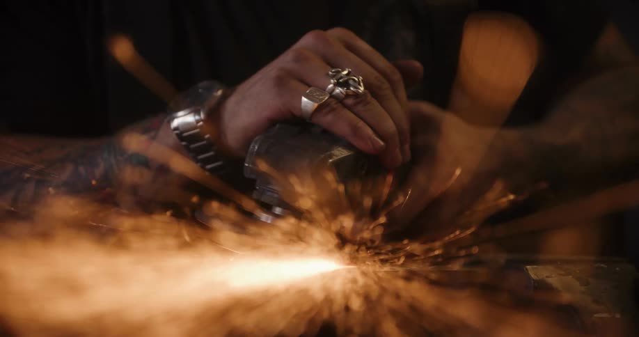 A tattooed craftsman uses an angle grinder, sending a cascade of bright sparks flying from the metal in a workshop