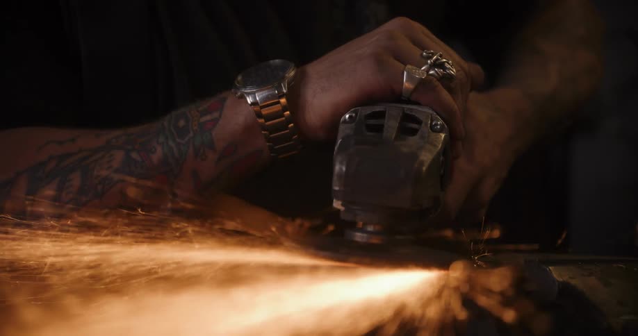 Slow-motion close-up of a craftsman's tattooed hands grinding metal, creating a brilliant shower of sparks