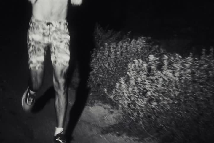 A shirtless man wearing a headlamp runs toward the camera at night on a dark trail in grainy black and white